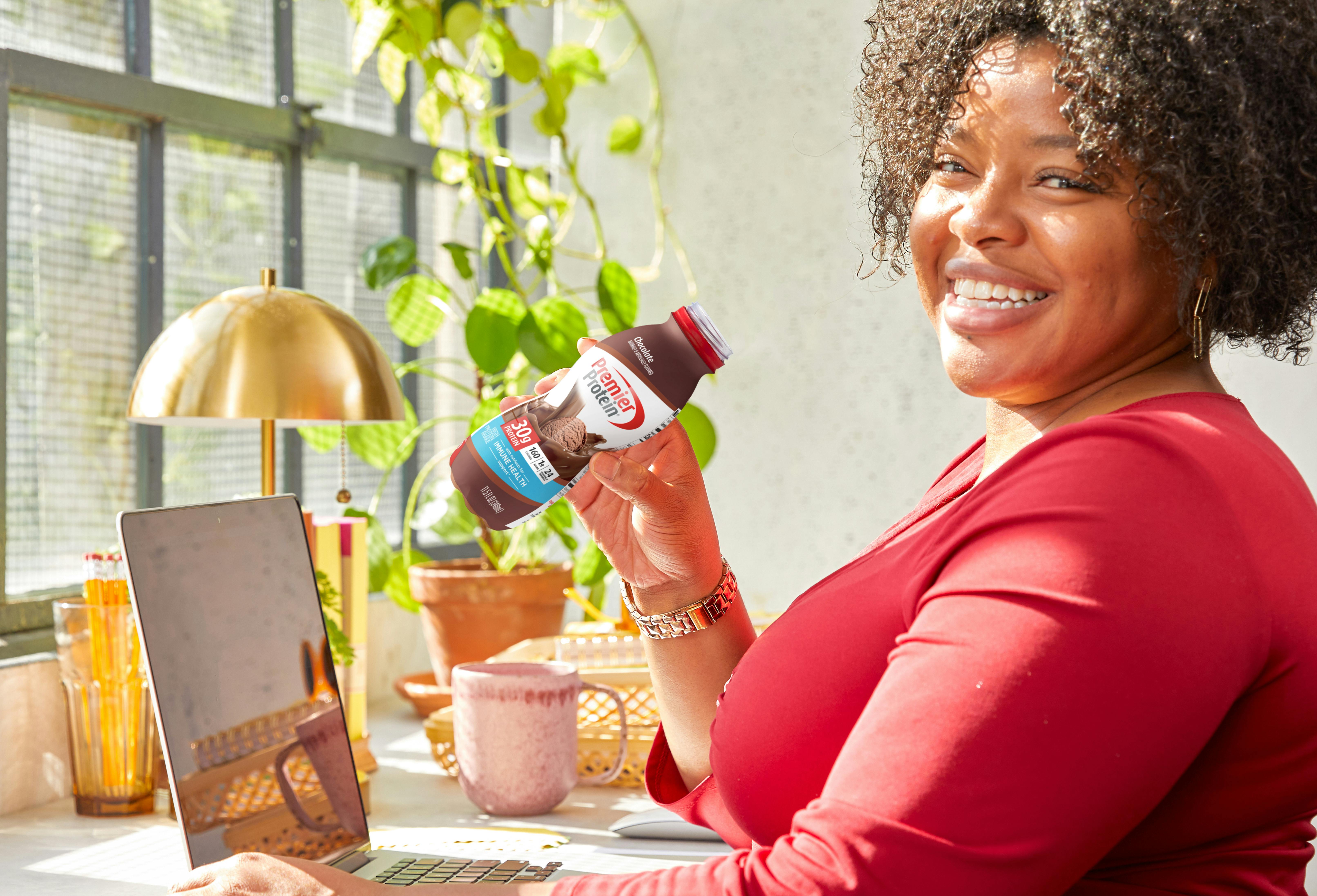 A smiling woman drinking a premier protein chocolate shake while working on a laptop