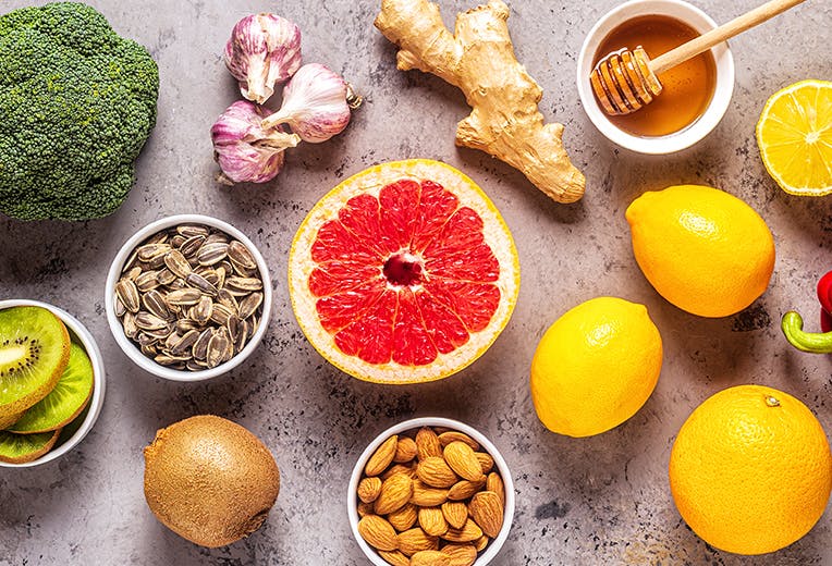 Top-down shot of a countertop with broccoli, lemons, grapefruit, ginger, red pepper, kiwi, sunflower seeds, honey, and almonds.