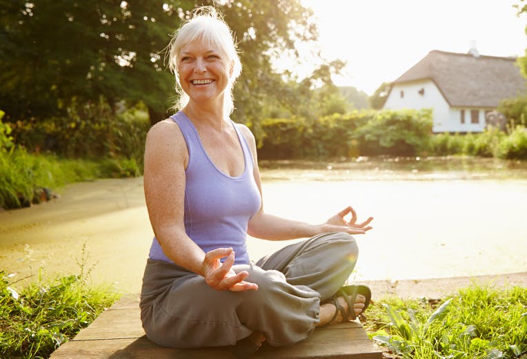 A woman meditating in the sunshine next to a small pond.